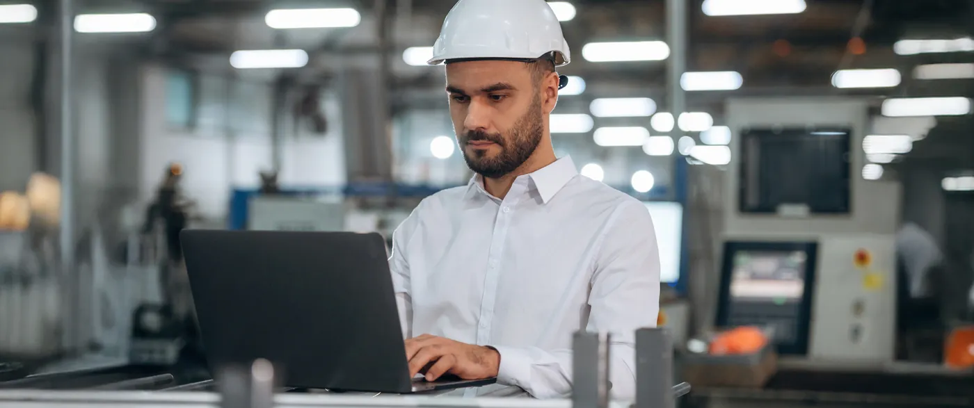 Engenheiro de capacete branco pesquisando num notebook dentro de uma fábrica