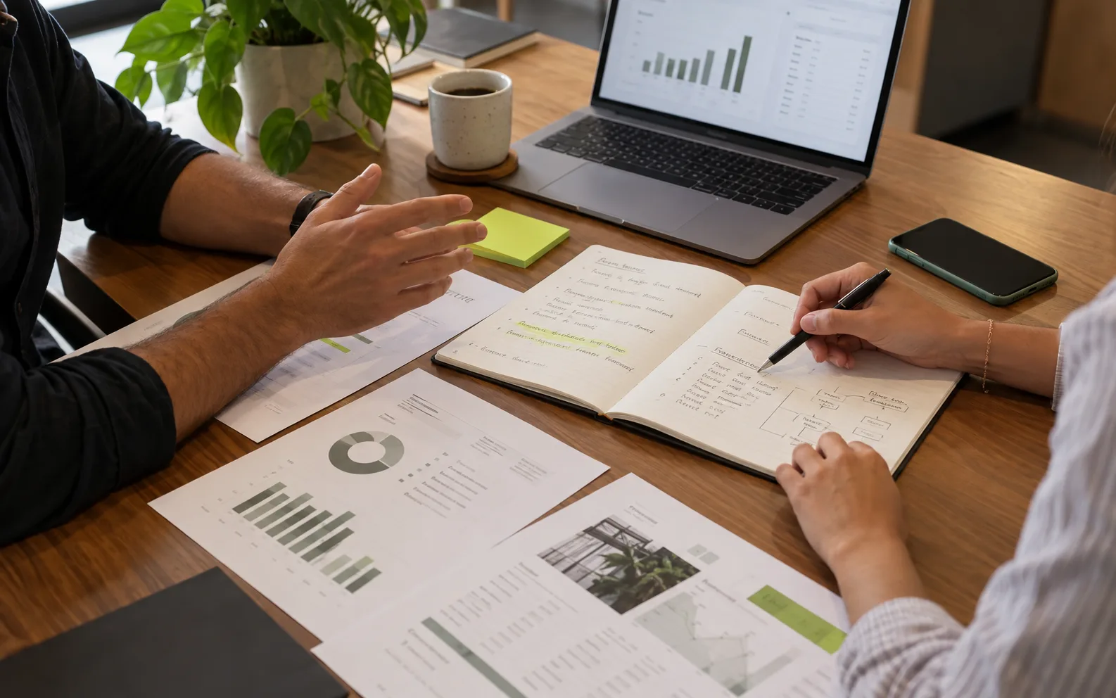 Duas pessoas à mesa em uma conversa de diagnóstico, com anotações no caderno, relatórios impressos e um gráfico no notebook